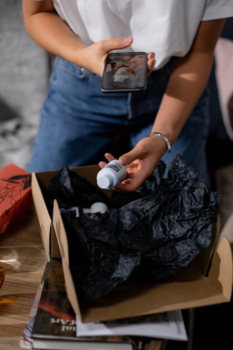A woman examines a bottle of cosmetics from an online order, capturing the moment on her smartphone.