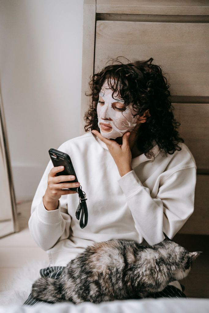 Calm female in facial moisturizing mask sitting near wardrobe with cat on laps while browsing cellphone in room during weekend